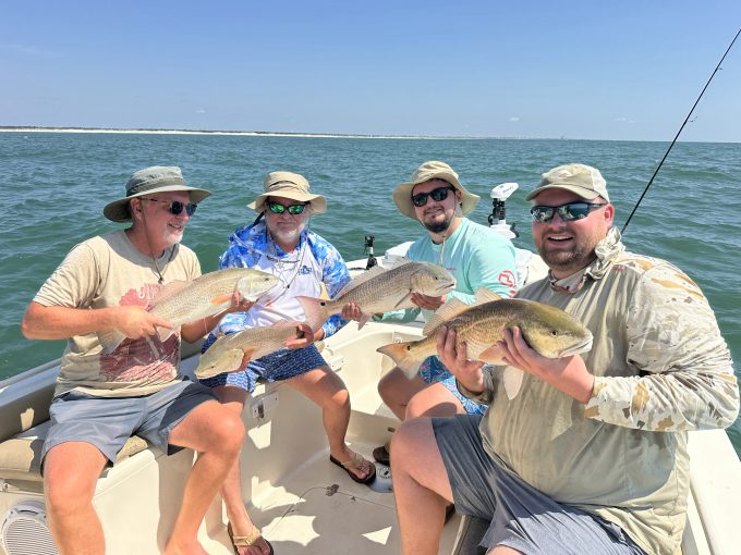 20260906_154855354_iOS Four anglers each holding a redfish aboard a fishing boat, with a sandy shoreline in the background.