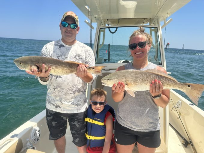 myrtle-beach-red-drum-fishing-with-kids Two adults holding large red drum with a child wearing a life jacket between them aboard a charter boat.