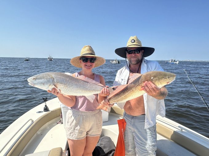 north-myrtle-beach-bull-red-drum-charter-catch Two anglers wearing straw hats and holding large bull red drum, with fishing boats in the background.
