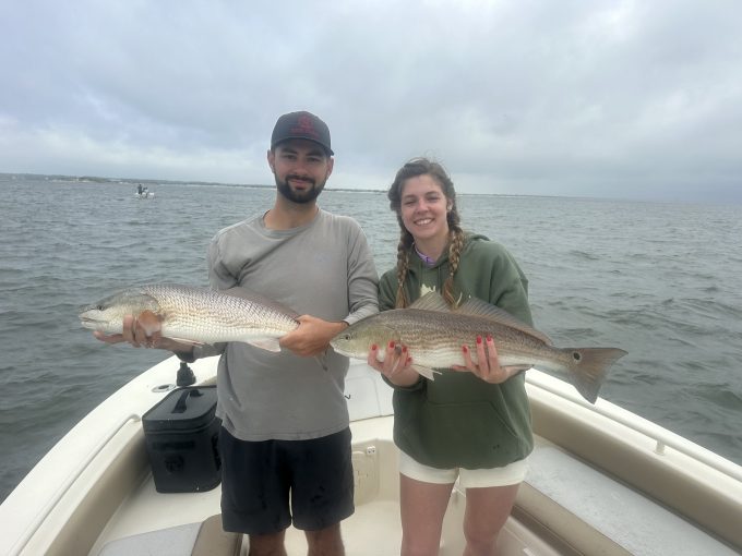 north-myrtle-beach-redfish-charter-anglers Two anglers holding redfish horizontally aboard a fishing boat under an overcast sky.